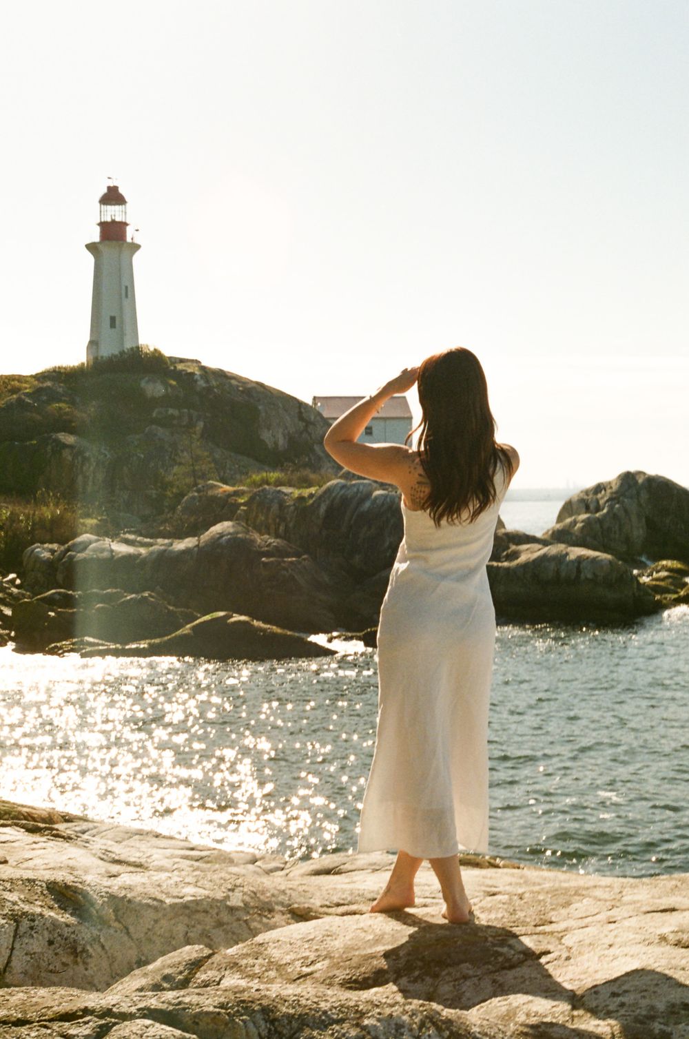 Figure stands on rocky shore near lighthouse during sunset with ocean waves glittering.