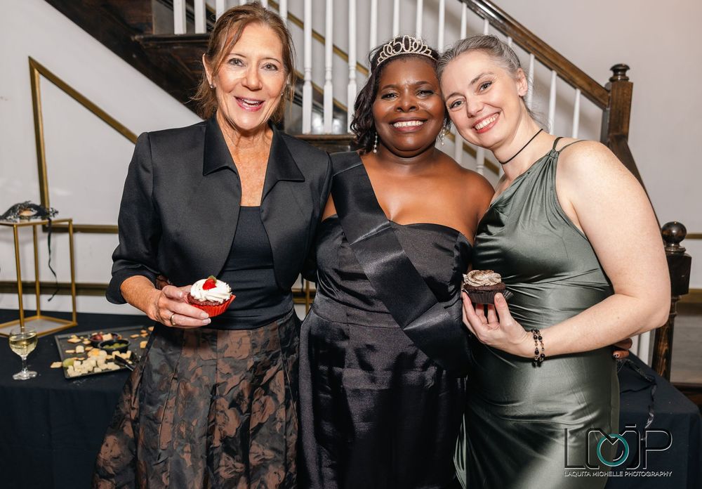 Three women in elegant evening attire smile together at a formal event.