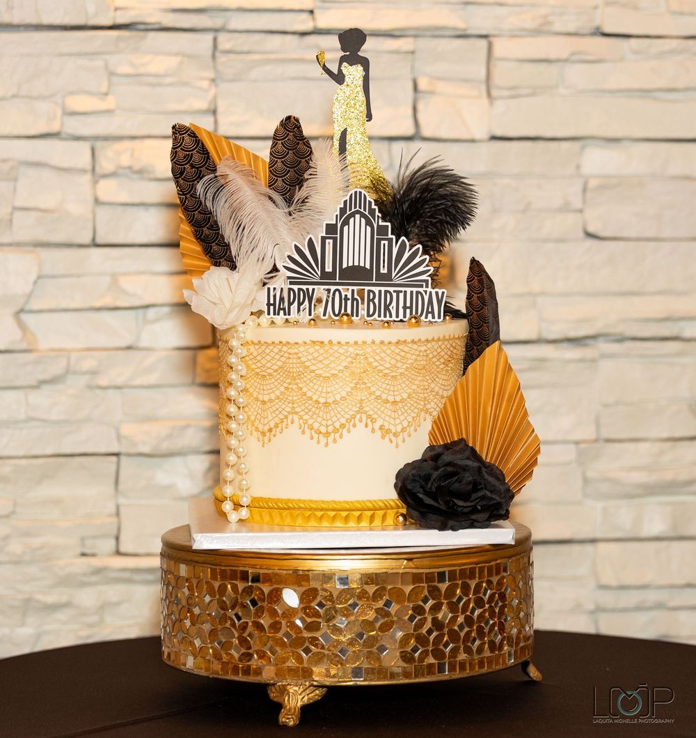 Elegant black and gold themed birthday cake with lace details and feather decorations on a white brick background.
