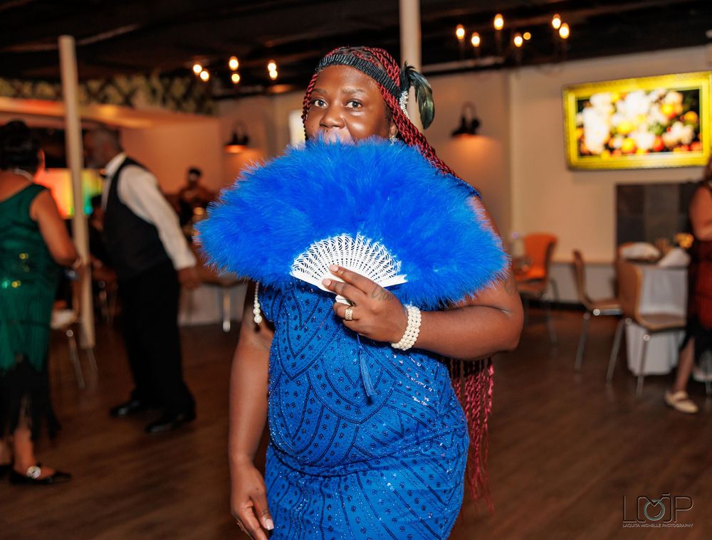 A guest in a vibrant blue dress holds a matching blue feather fan while enjoying a festive indoor event.