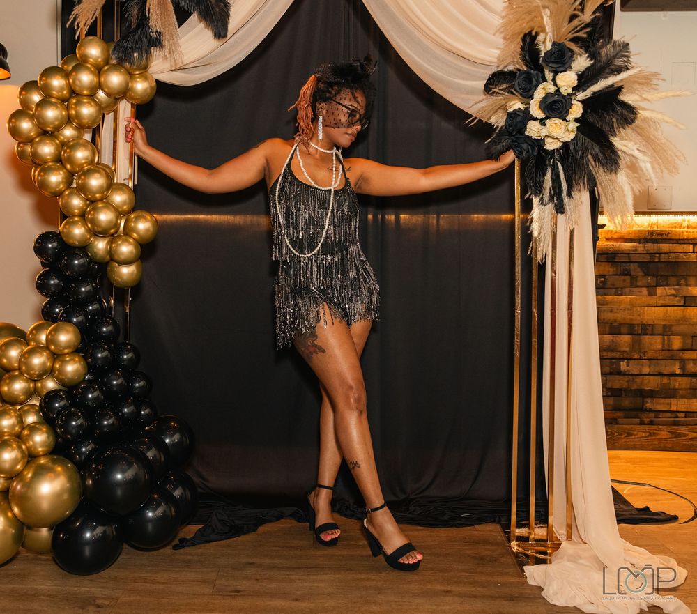 A 1920s-themed party setup with gold and black balloons, white drapes, and elegant decor against a brick wall backdrop.