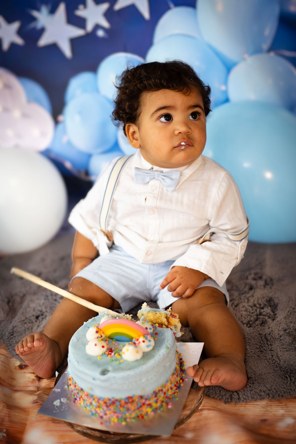 Baby in formal outfit sitting beside rainbow-decorated birthday cake with star backdrop.