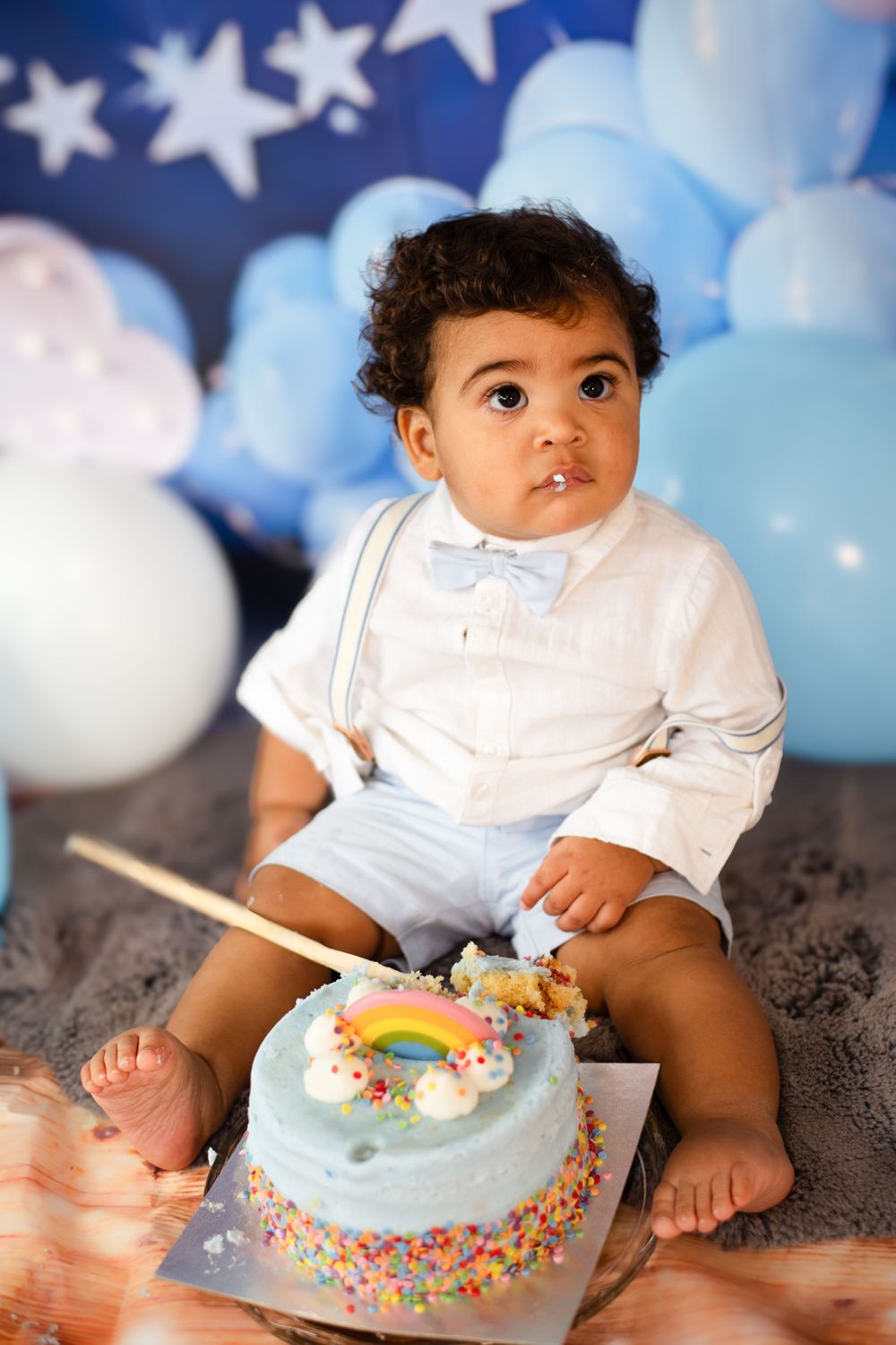 Baby in white button-down shirt sitting with blue birthday cake decorated with rainbow design.