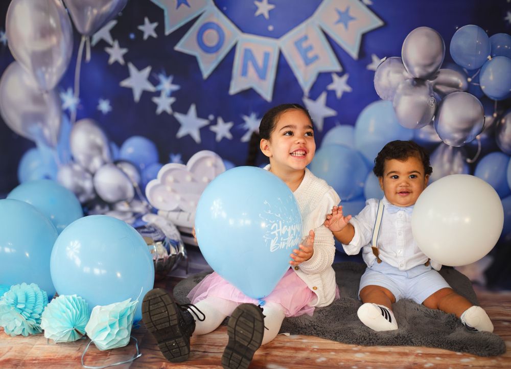 Two children playing with blue and white balloons against decorated backdrop at birthday celebration.