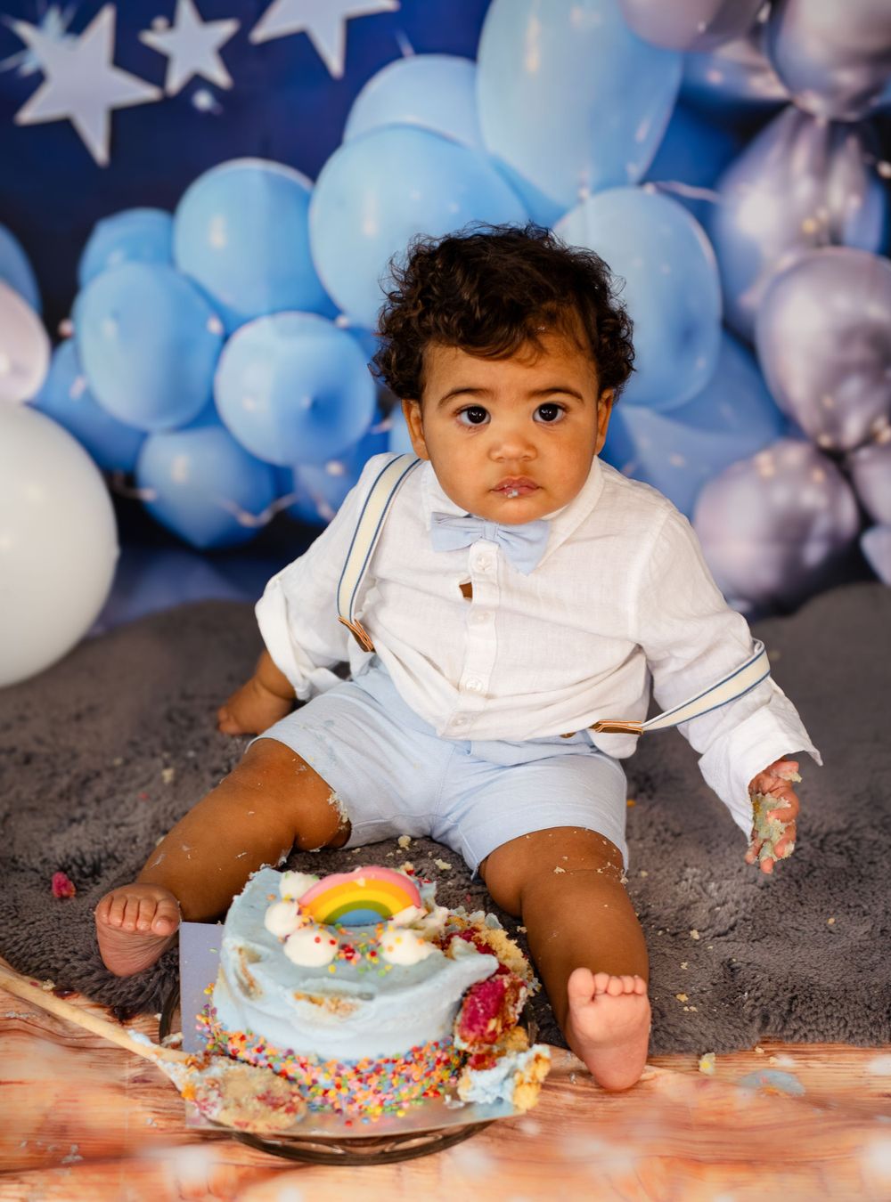 A baby in light blue outfit sits by a smashed birthday cake with balloon decorations.