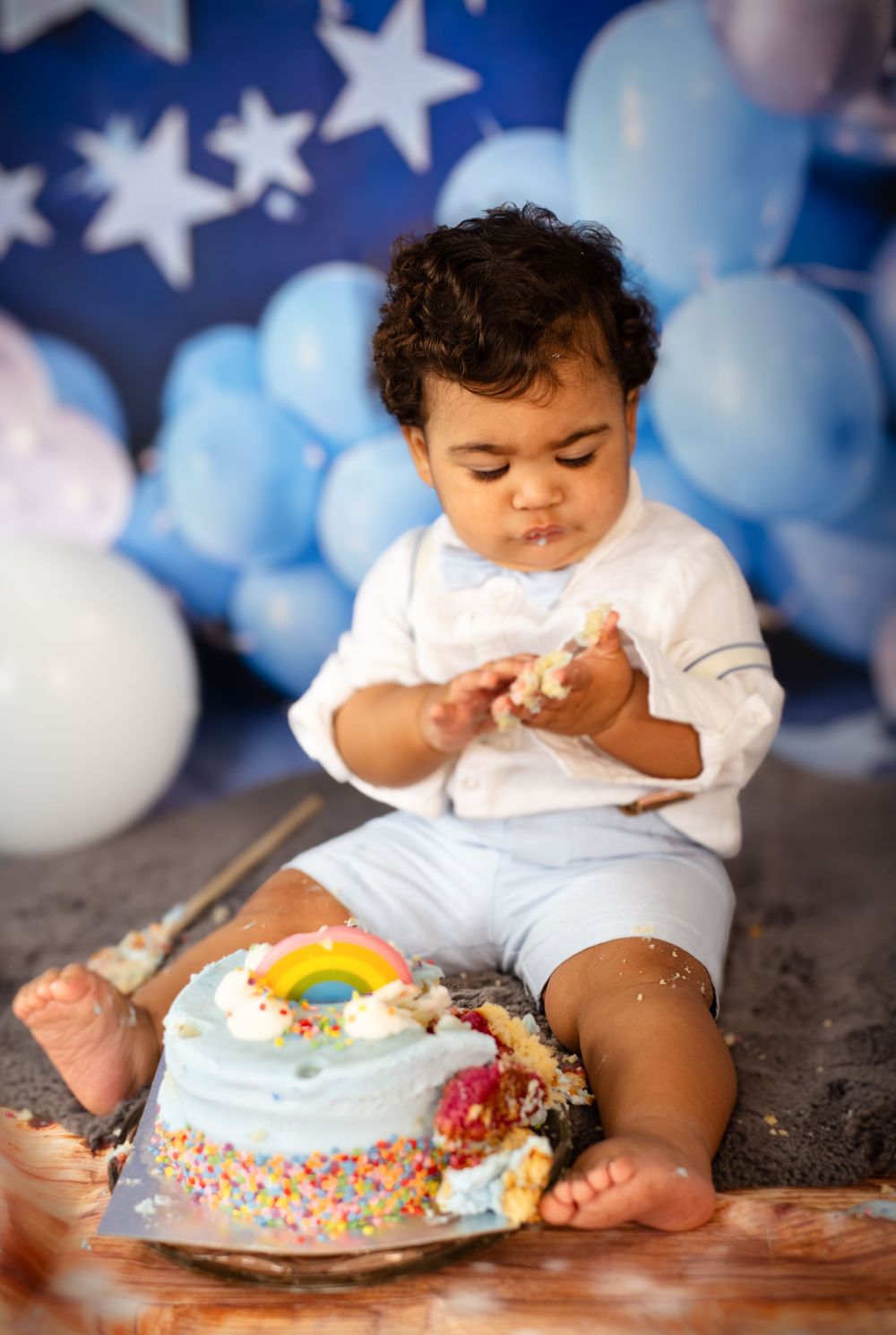 Baby examining cake during first birthday photoshoot with blue and white balloon decorations.
