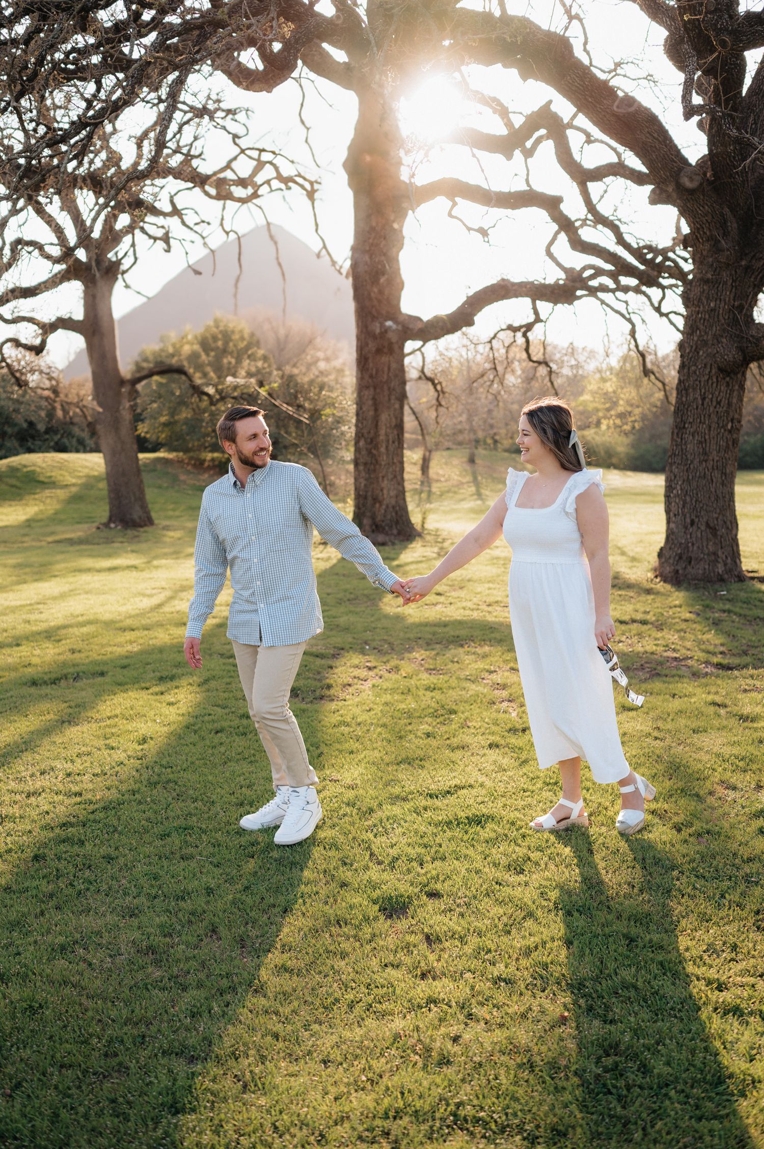 A couple holds hands while walking through a sun-dappled grove of trees in white and blue outfits.