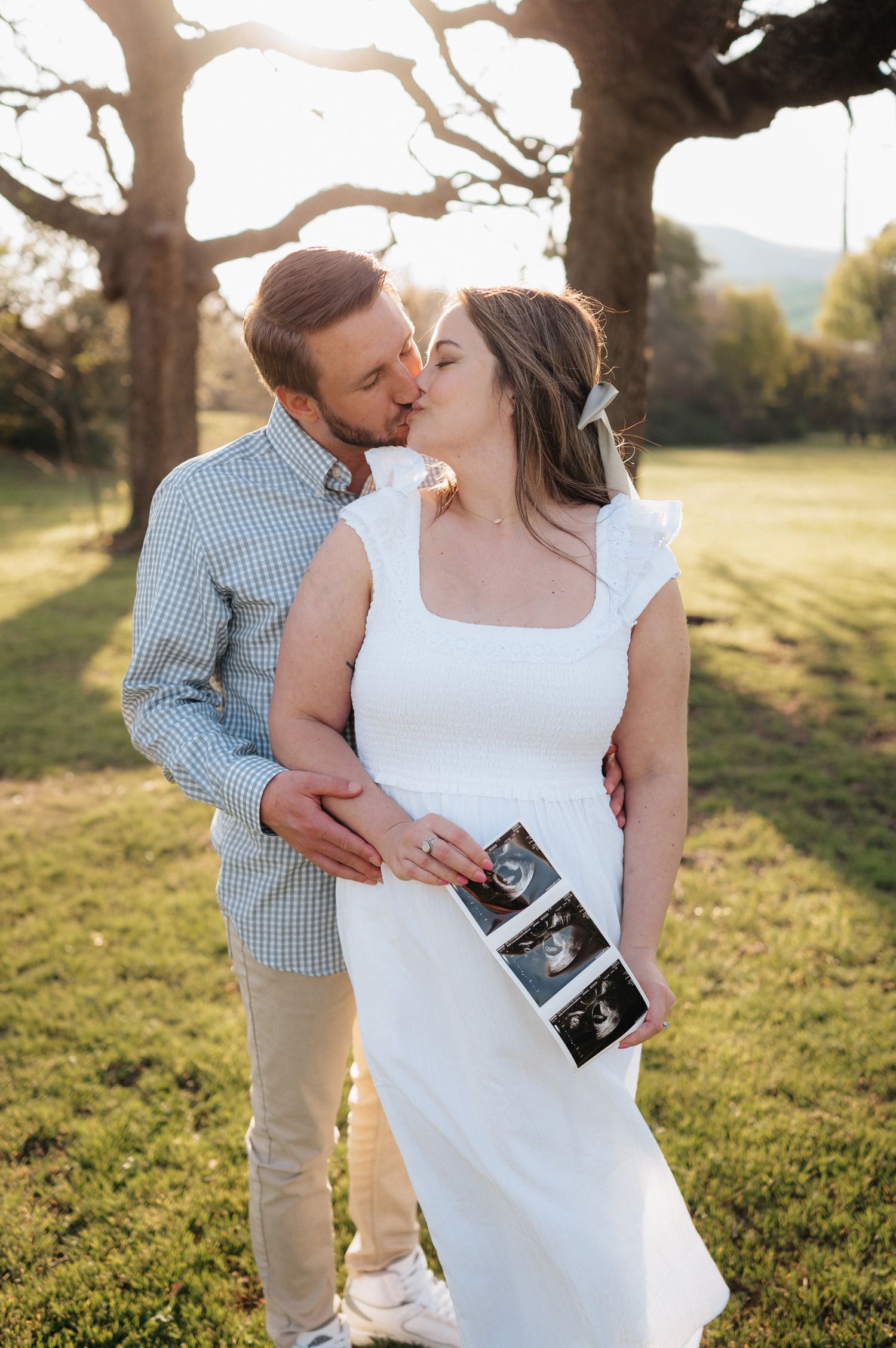 A couple shares an intimate moment outdoors while holding ultrasound photos in golden sunlight.