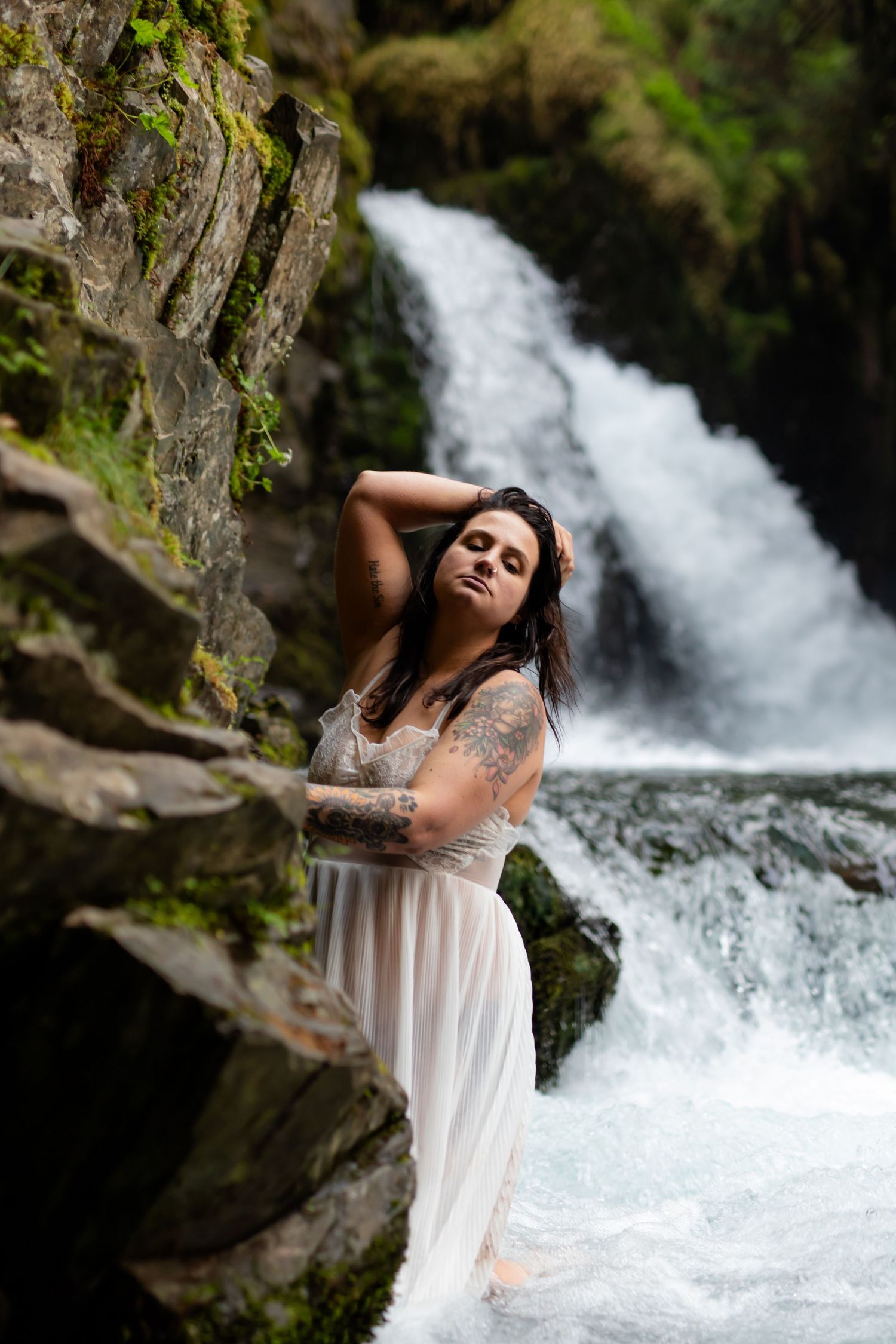 A woman in a flowing white dress poses dramatically by a rushing waterfall.