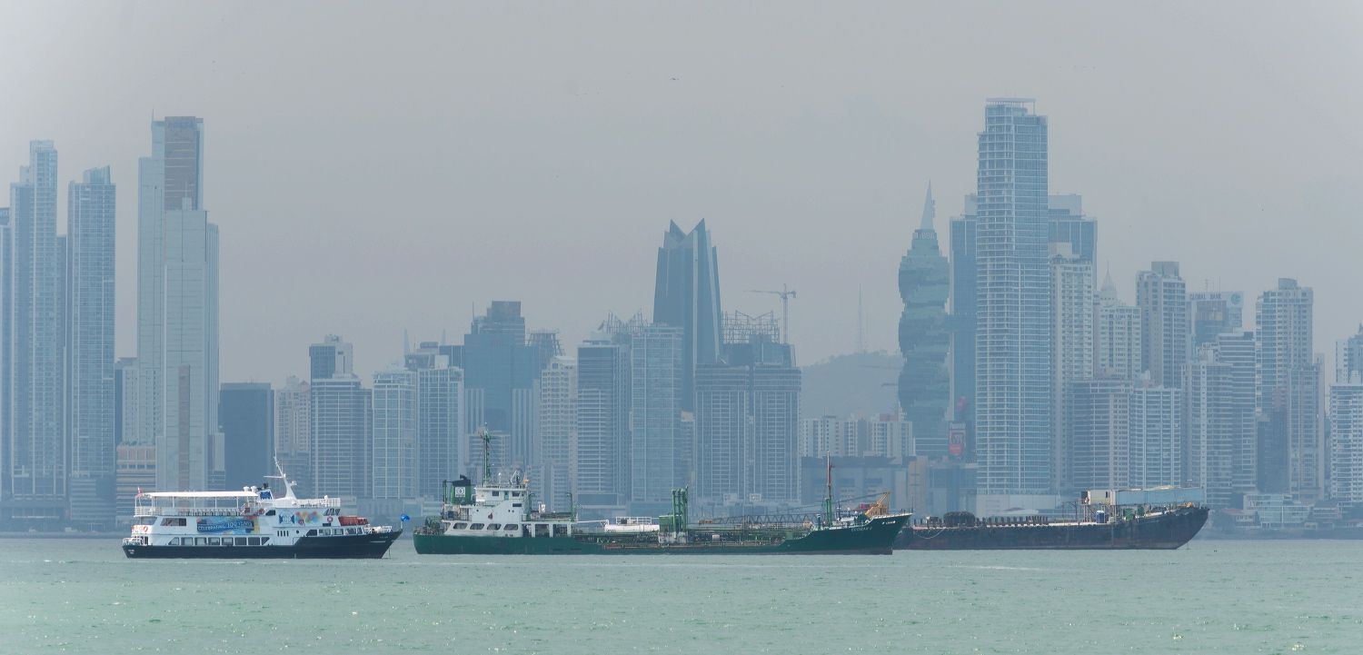 Container ships anchored in front of a hazy city skyline with modern skyscrapers.
