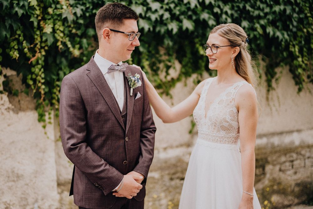 A tender moment between newlyweds wearing glasses as they share a happy exchange against an ivy-covered wall.