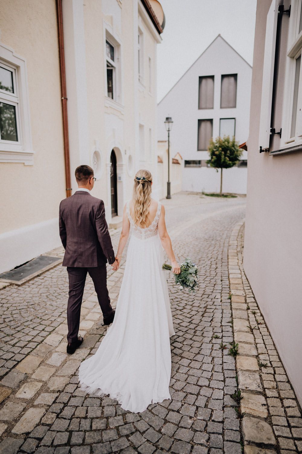 A couple walking hand in hand down a quaint cobblestone street between cream-colored buildings.