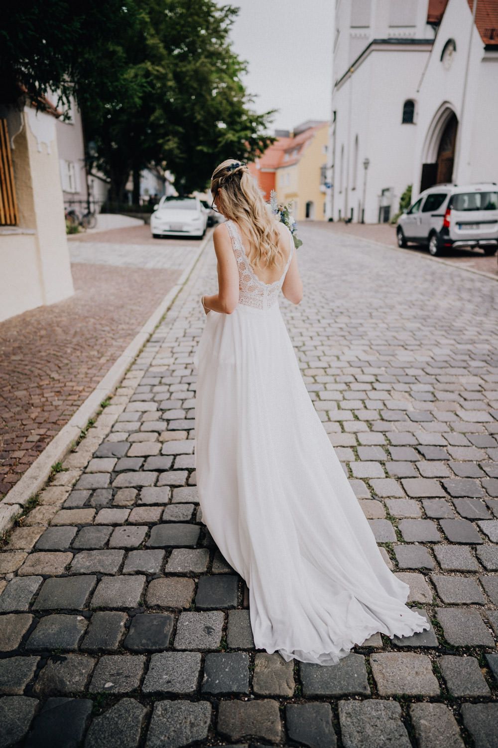 Flowing white wedding dress photographed from behind on cobblestone street in European town setting.