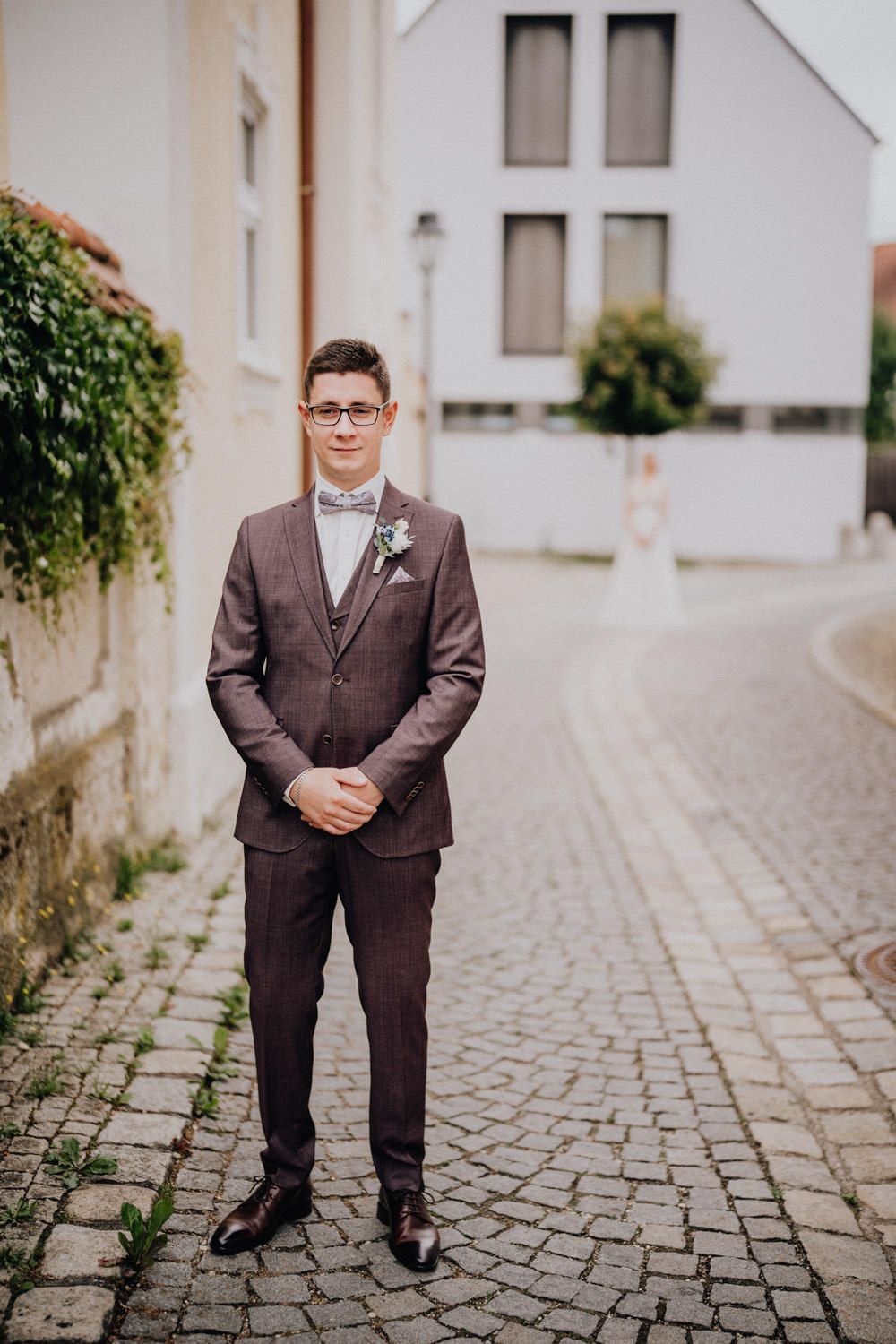 Groom in brown suit and bowtie standing on cobblestone street during wedding photoshoot.