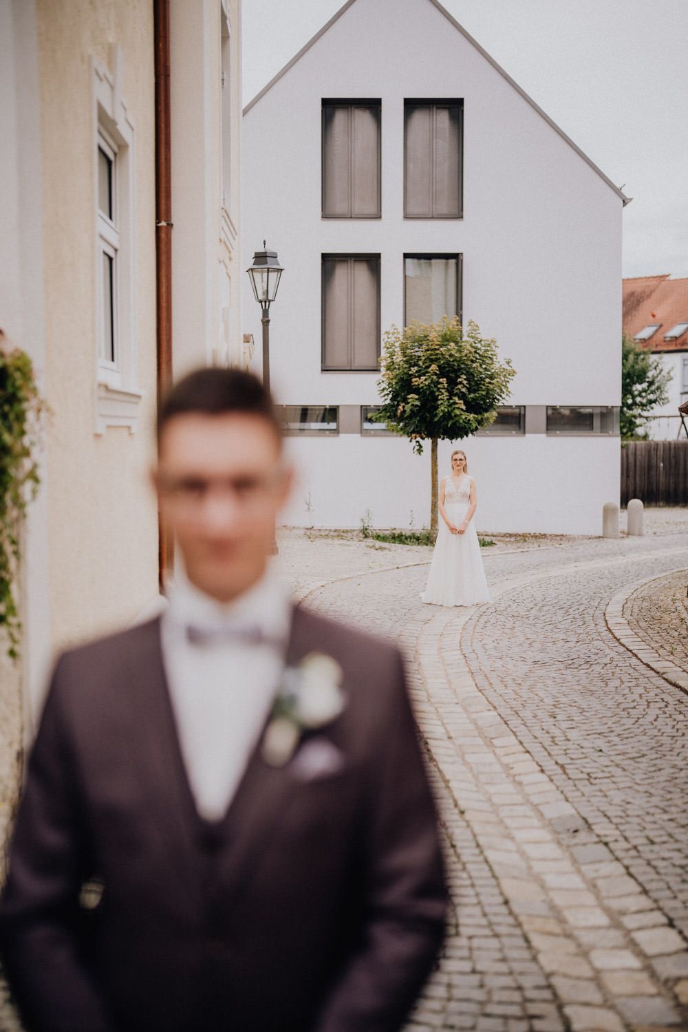 Wedding couple standing apart on cobblestone street with white building in background.
