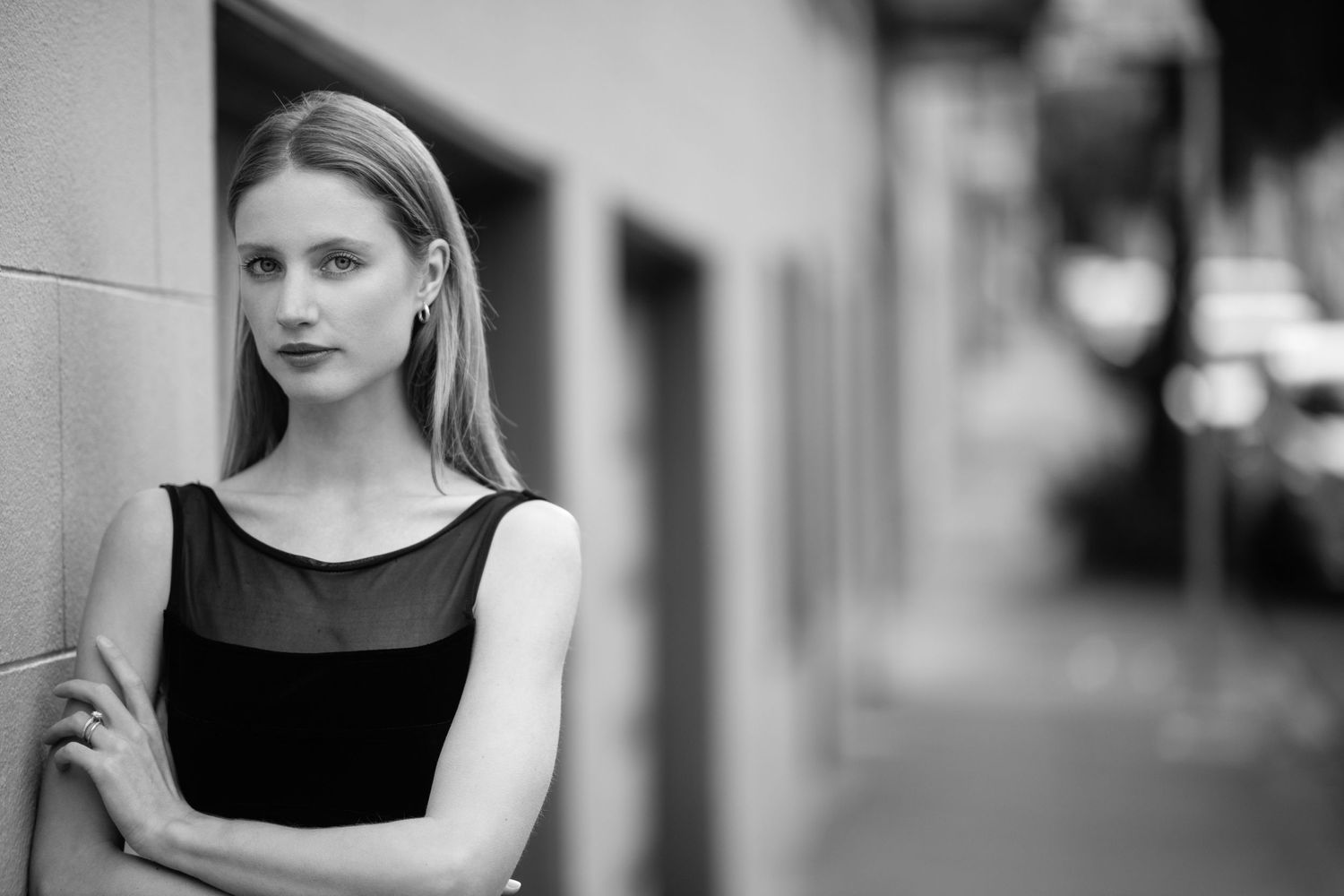 Black and white portrait of a person in a sleeveless dress standing against a wall with a soft blurred background.
