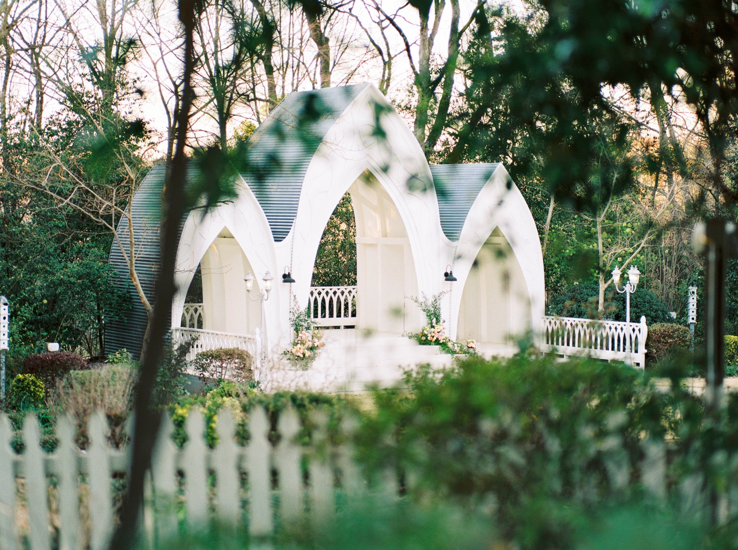 Whimsical white garden chapel with gothic arches nestled among trees.