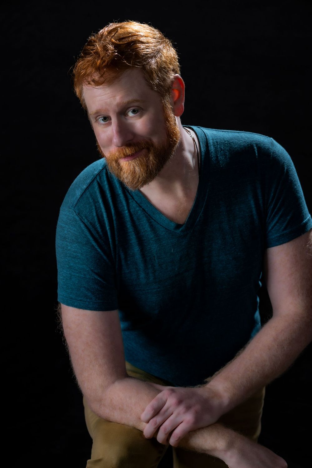 Portrait of a person with red hair and beard wearing a teal v-neck shirt against a dark background.