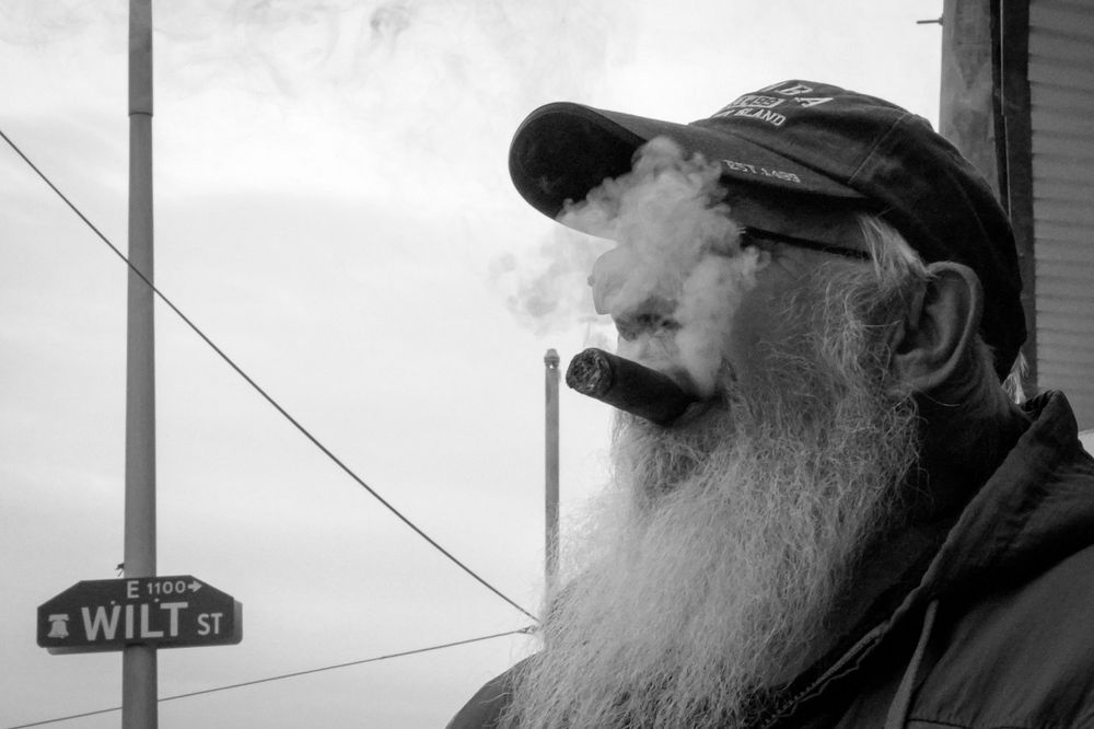 Black and white portrait of a bearded person smoking with vapor visible against a cloudy sky.