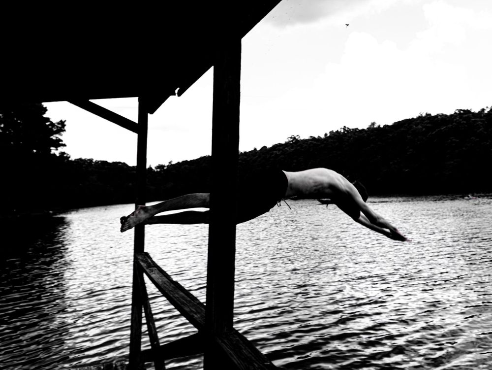 Silhouette of person diving into lake from wooden platform at sunset.