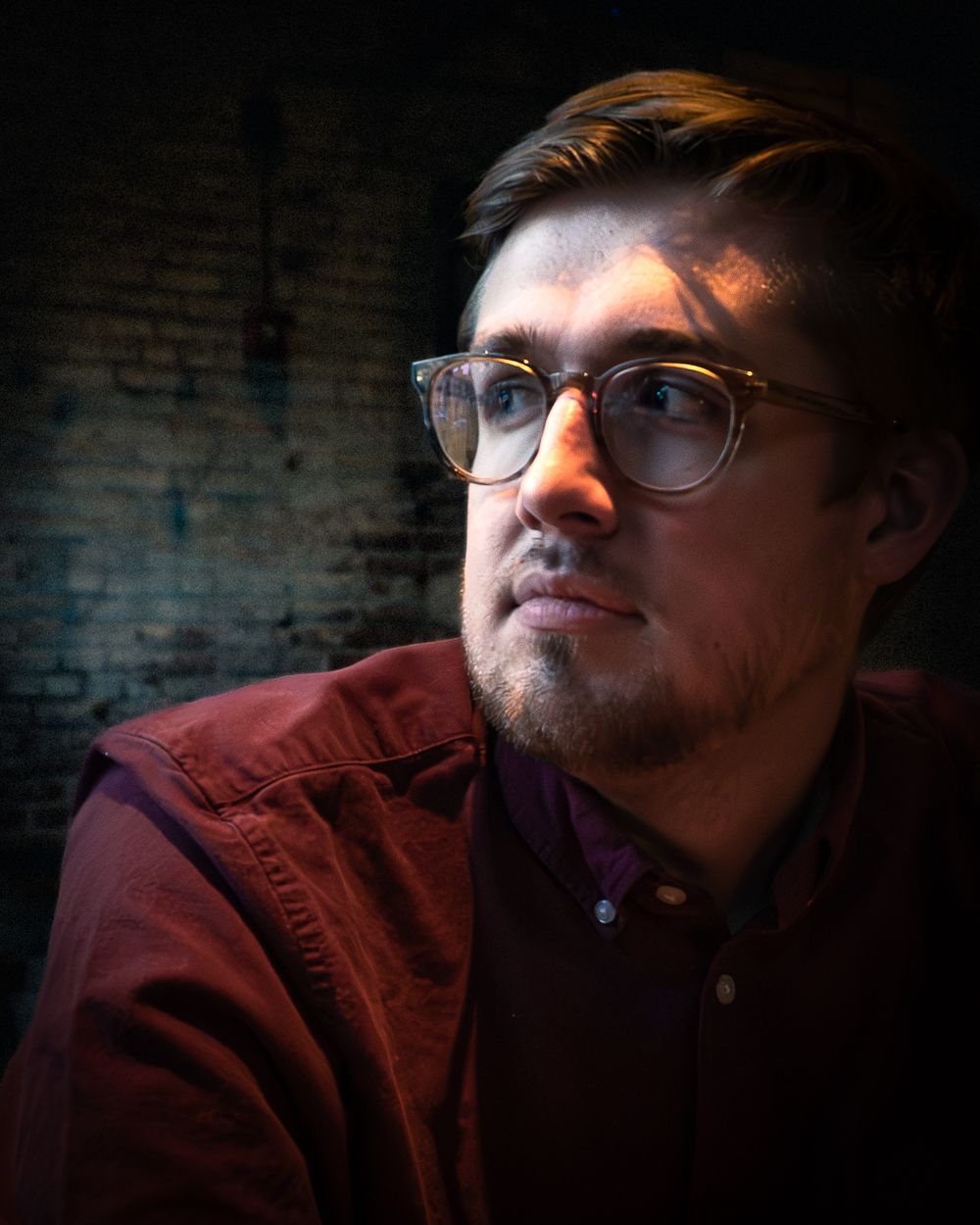 Dramatic portrait of a person in glasses and maroon jacket against brick wall with moody lighting.