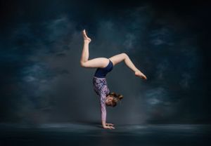 A gymnast performs a one-handed handstand in a dramatic pose against a dark, misty background.