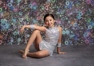 Young girl in silver sequin dance costume poses gracefully on floor in front of colorful floral backdrop.