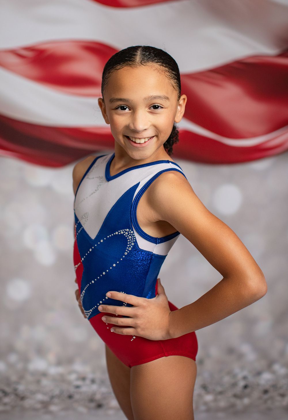Young gymnast in red white and blue leotard poses against American flag background.