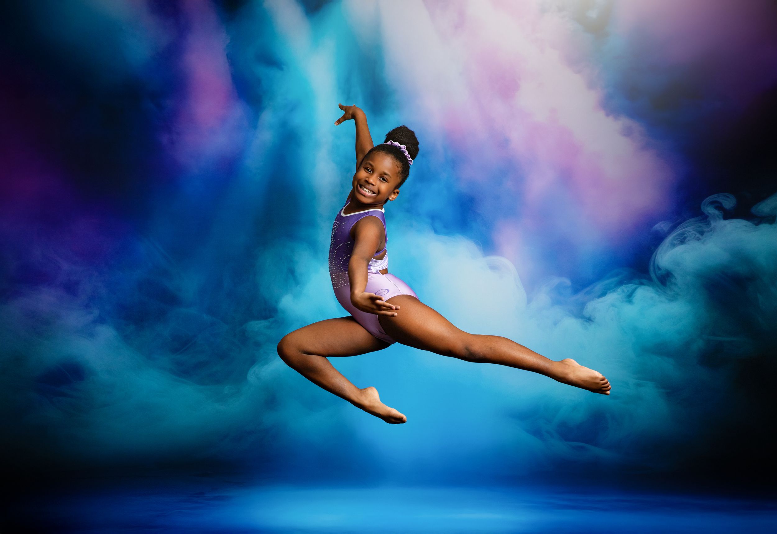 A dancer in a turquoise leotard leaps gracefully through the air against a dreamy blue and purple clouded backdrop.