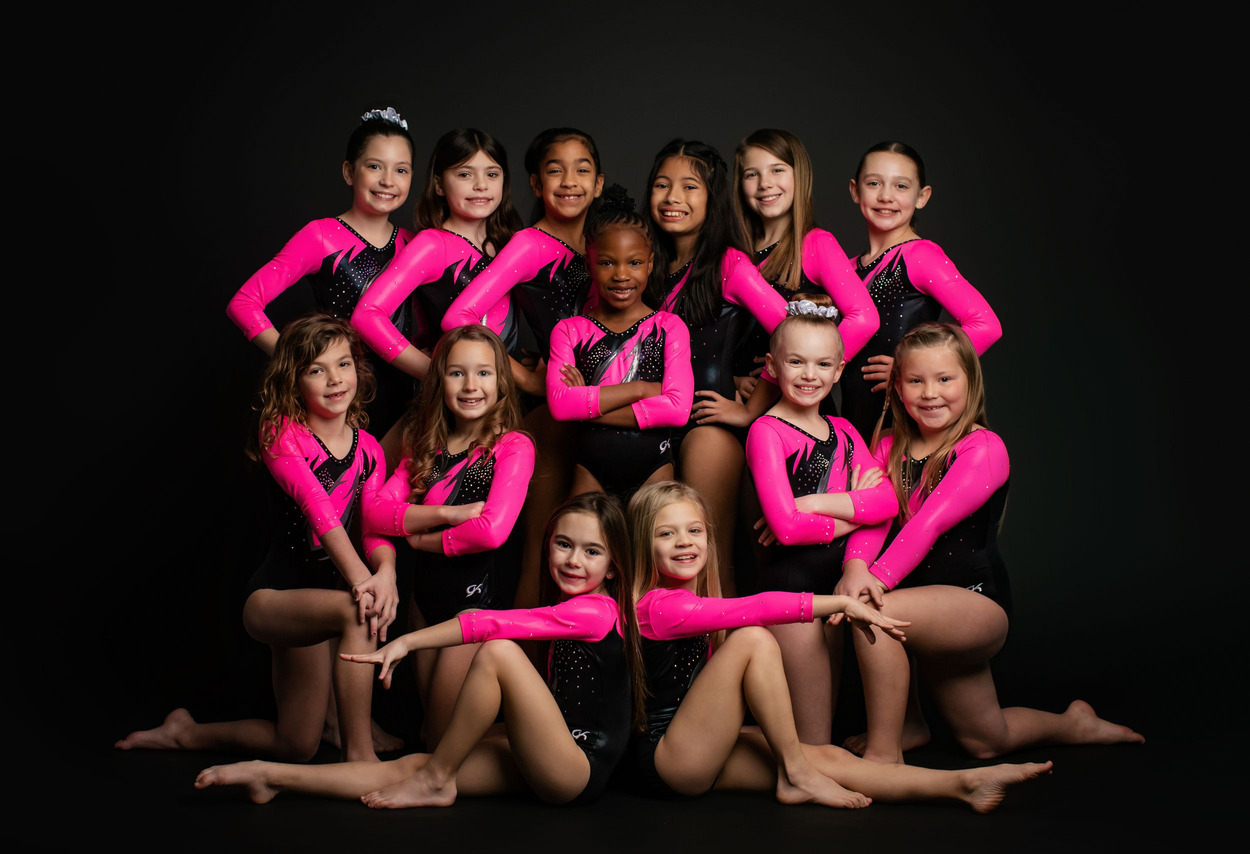 Group of gymnasts in black leotards pose together against dark background in artistic formation.