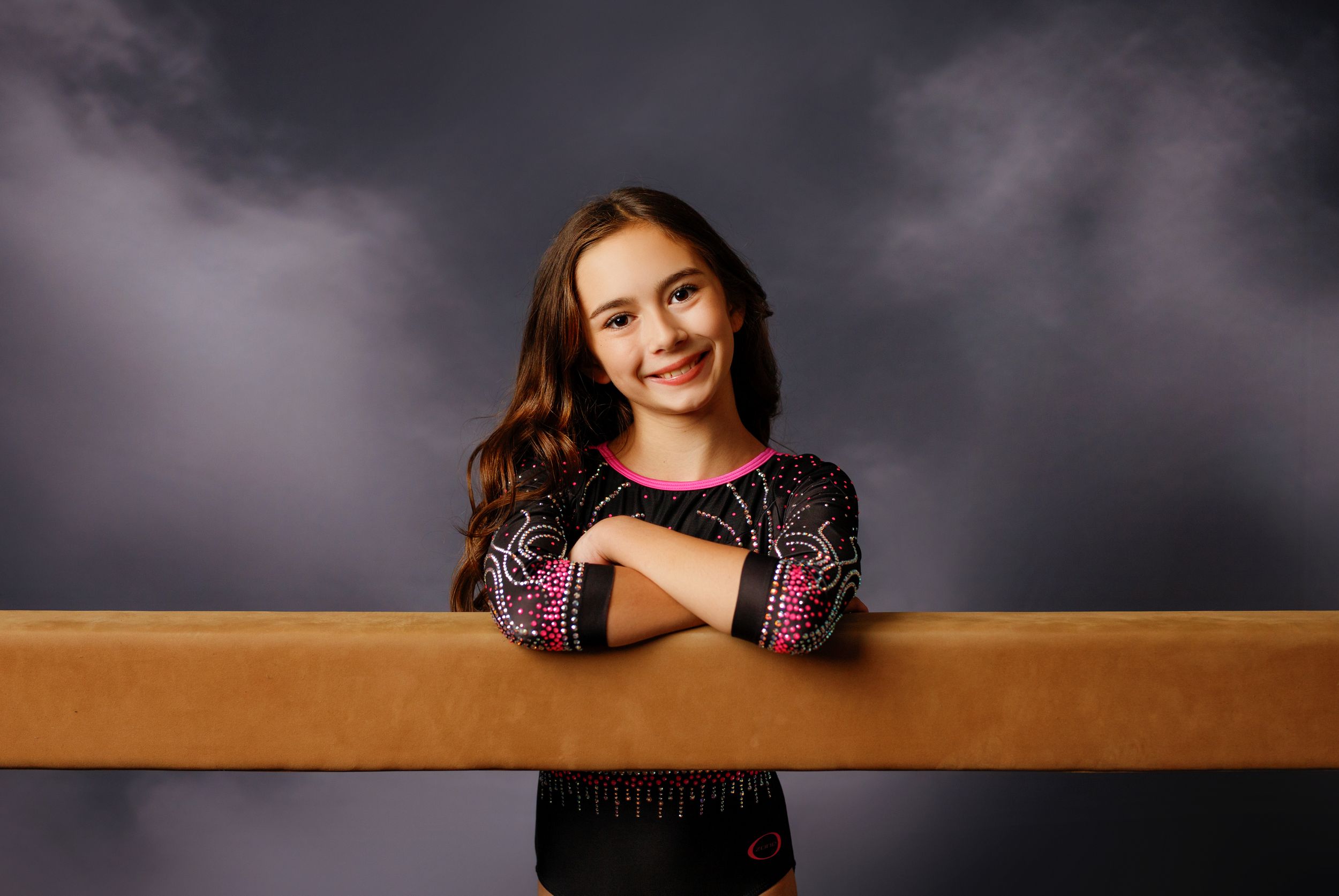 Young gymnast in decorated leotard smiling while leaning on a balance beam against a dark studio backdrop.