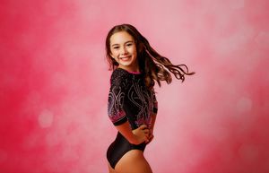 Young gymnast smiling in black and pink rhinestone leotard posing against a vibrant pink bokeh background.