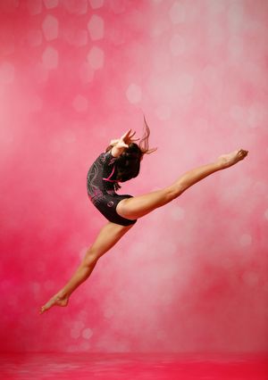 A gymnast in a black leotard leaps gracefully against a vibrant pink bokeh background.
