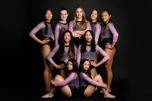 A gymnastics team poses together in purple leotards against a black background in a studio portrait.