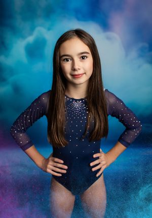 A gymnast in a navy blue sparkly leotard poses against a dreamy blue and purple clouded backdrop.