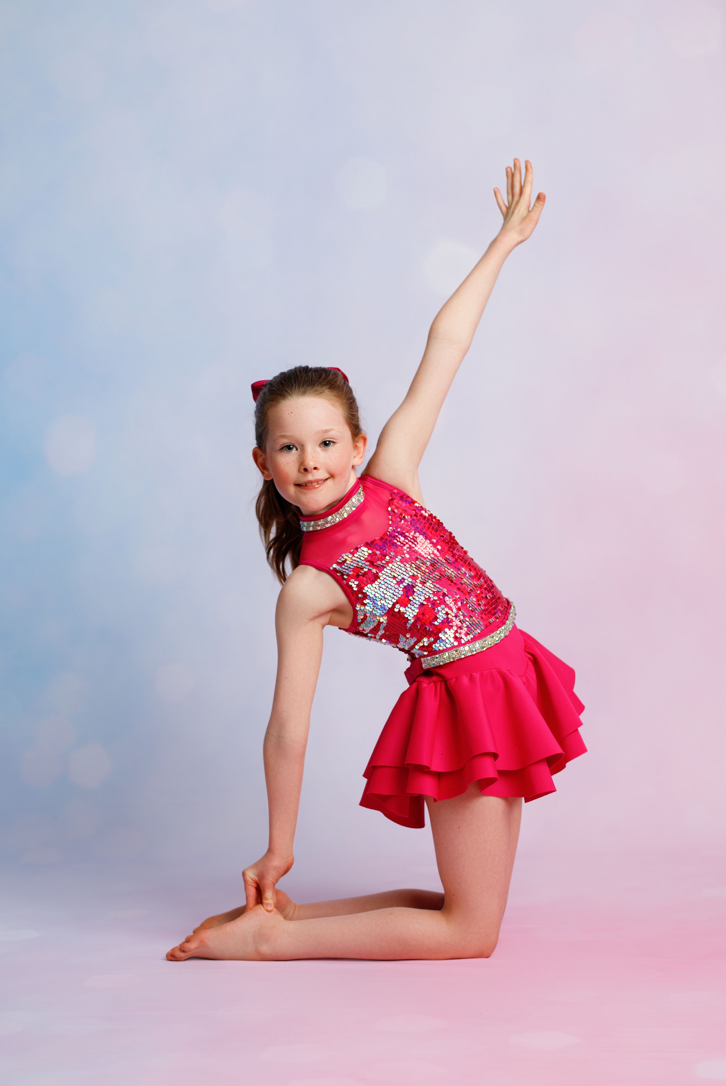 A young girl in a pink sequined dance costume poses gracefully on one knee with arm raised in a studio.