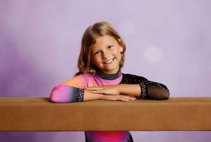 Young girl gymnast in sparkly pink and black leotard smiling while resting arms on a balance beam.