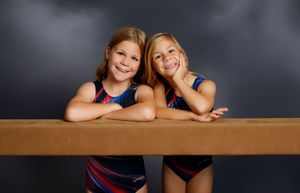 Two young girls in gymnastics leotards smiling and posing together at a balance beam against a dark background.