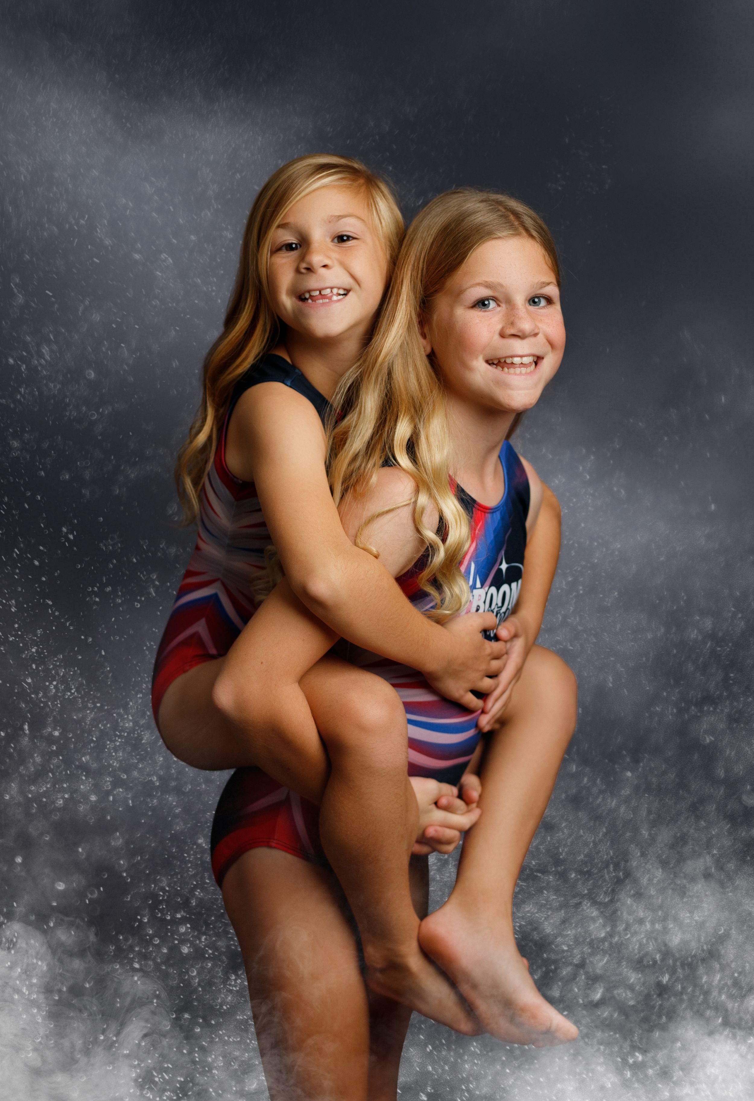 Two young girls in gymnastics leotards pose together, one giving the other a piggyback ride, smiling at the camera.