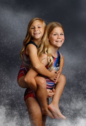 Two young girls in gymnastics leotards pose together, one giving the other a piggyback ride, smiling at the camera.