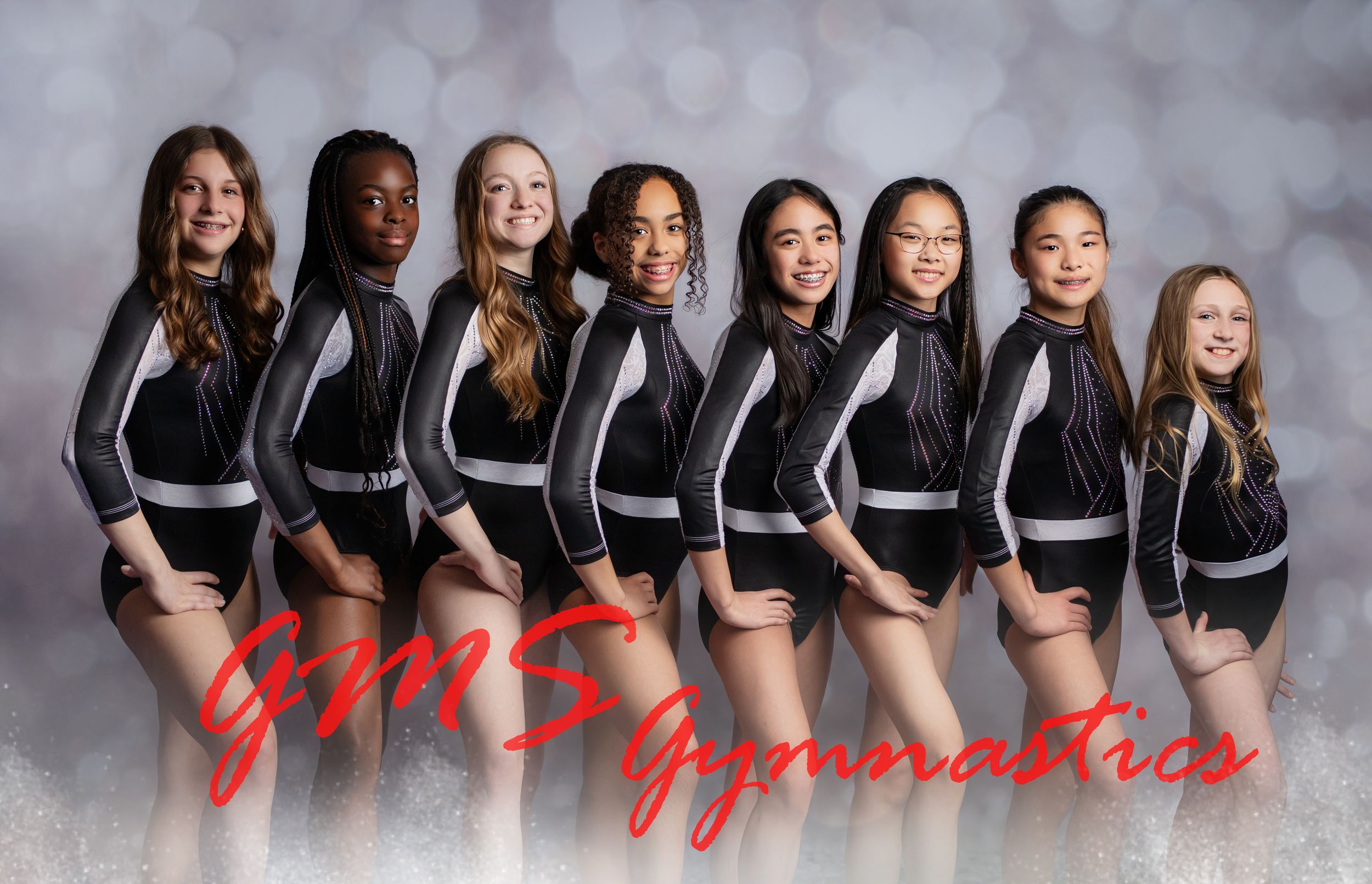 A group of dancers in black outfits pose together in a studio with a grey backdrop.