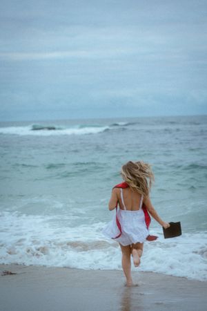 Person in white dress running through ocean waves while holding a dark folder.