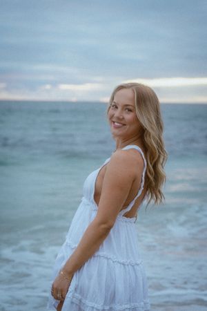 Person in white sundress posing playfully by the ocean waves.