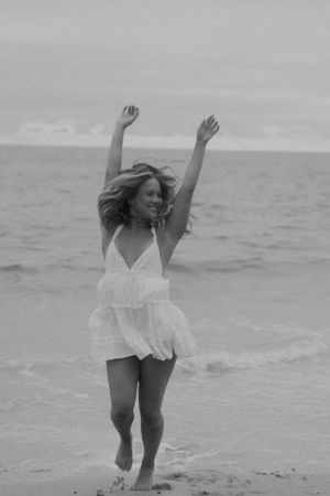 Black and white photo of person in white dress dancing joyfully on the beach.