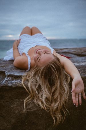 A person lying peacefully on dark rocks near the ocean at twilight.