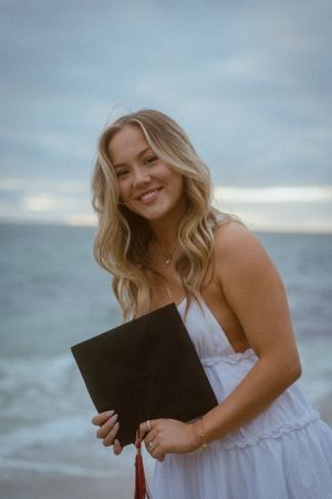 Person in white dress holding a black folder while posing at the beach.