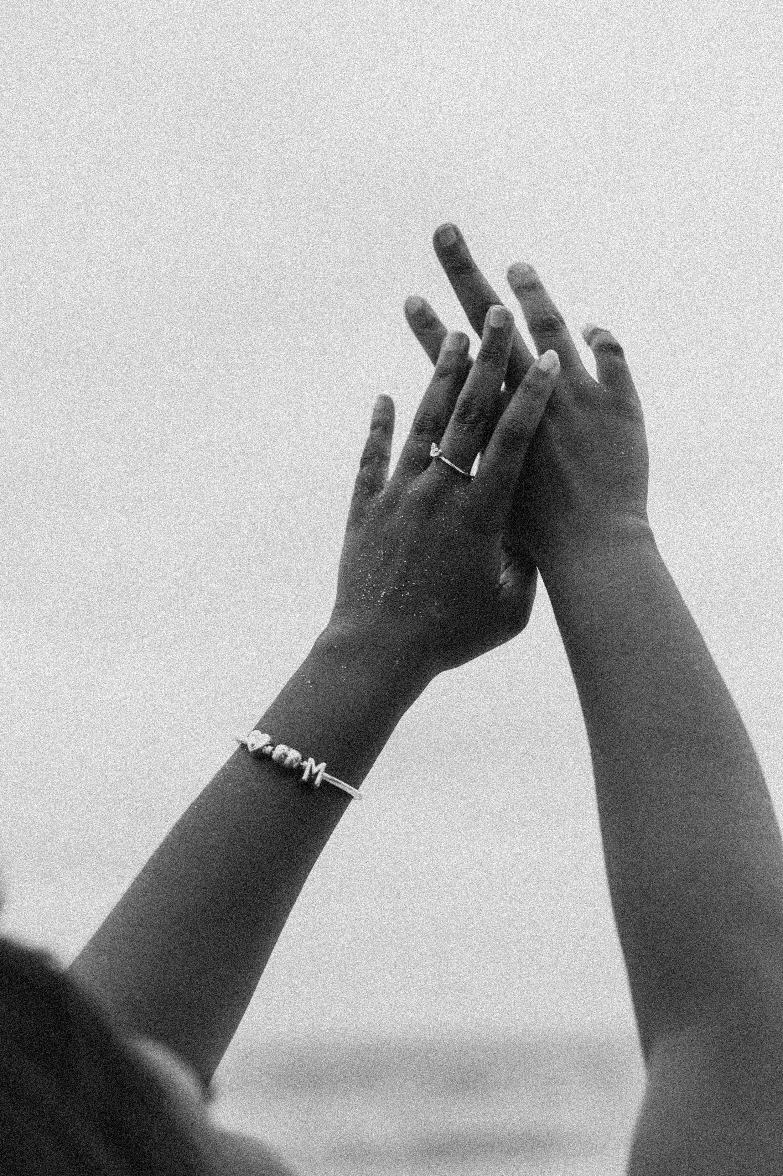Black and white photo of raised hands showing jewelry and rings against a bright sky.