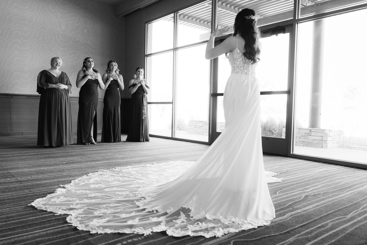 A wedding dress with a long lace train is showcased in front of large windows as bridesmaids look on in dark gowns.