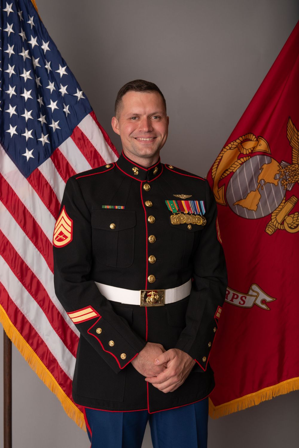 Marine in dress blues uniform stands at attention in front of American and Marine Corps flags for official portrait.