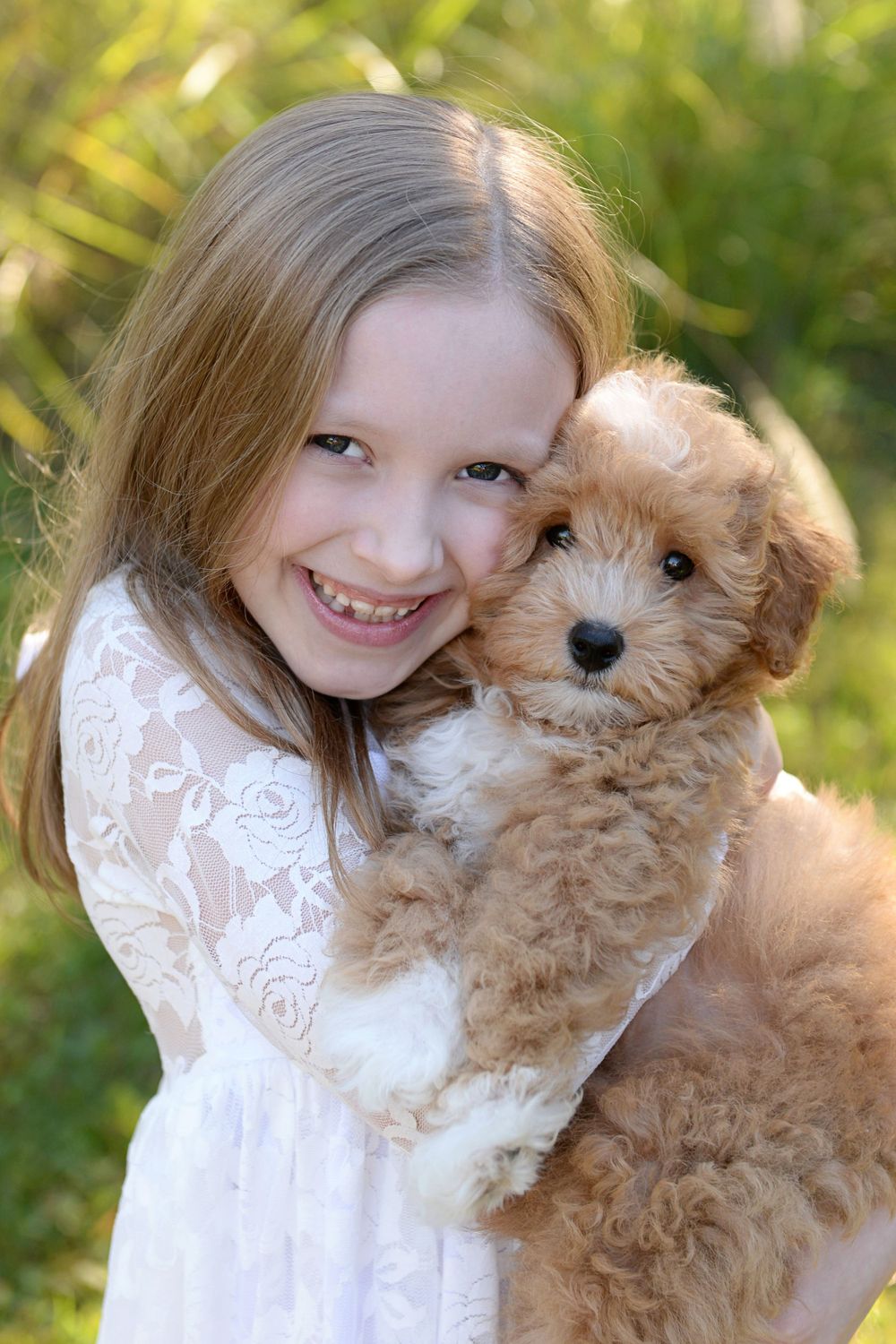 Young person in white lace dress hugging fluffy cream-colored puppy outdoors.